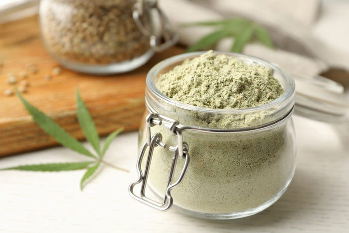 Glass jar filled with green powder on a light surface with cannabis leaves.
